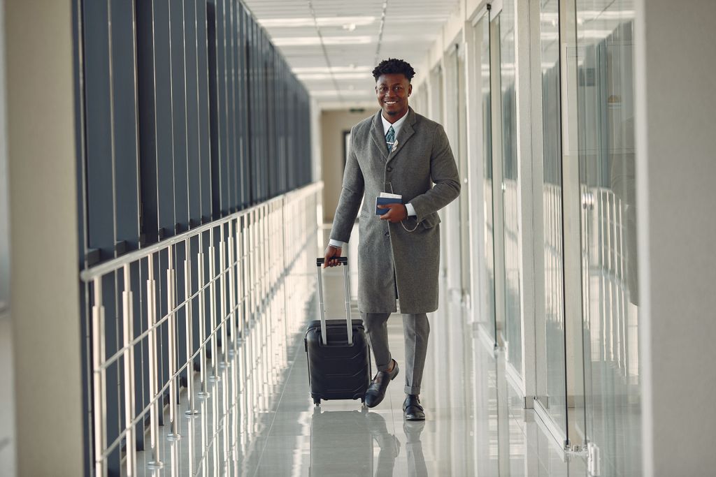 Full length of happy African American male in stylish classy gray clothes with suitcase and passport walking in contemporary airport terminal