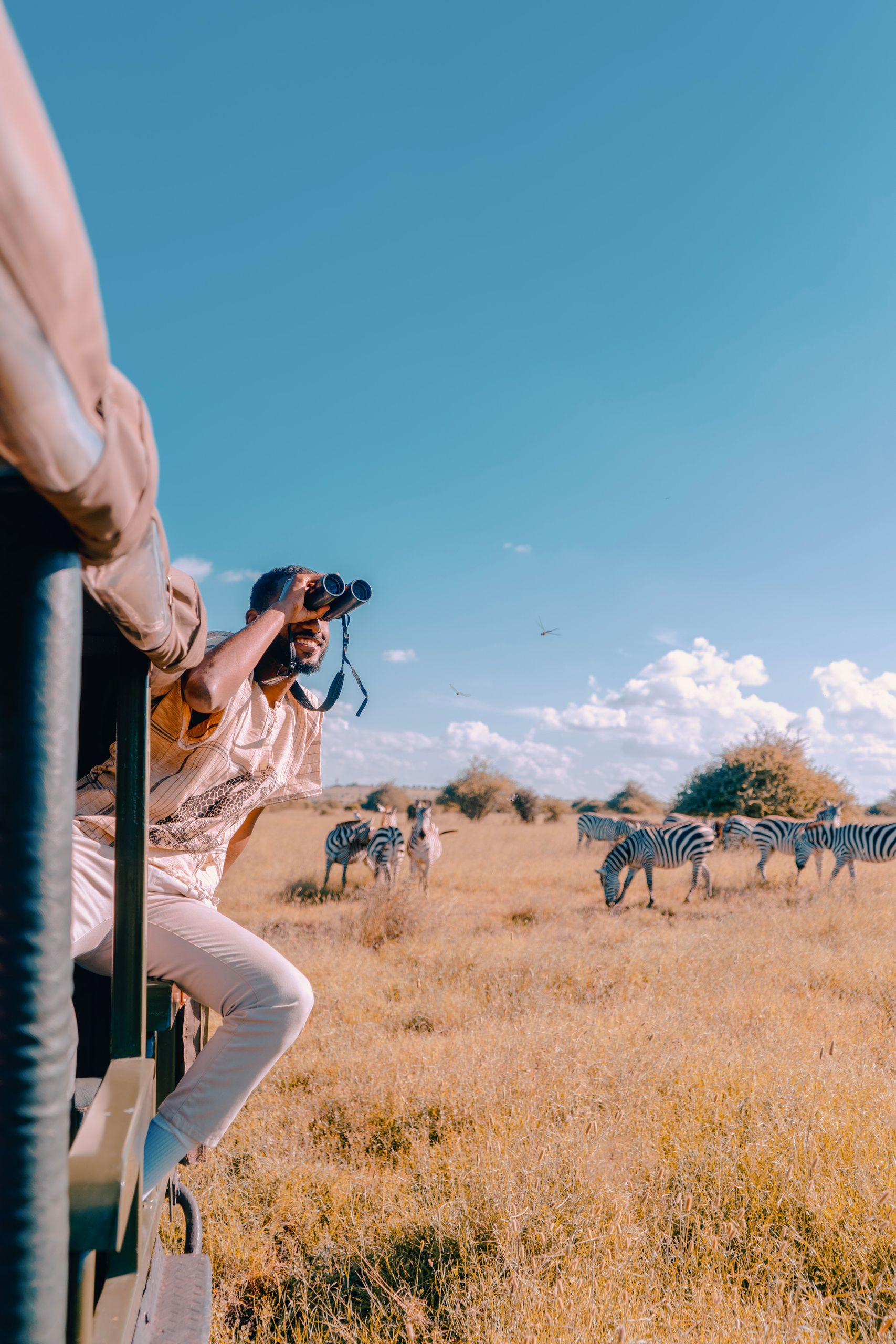 A man enjoys a wildlife safari in Nairobi, Kenya, observing zebras through binoculars from a truck.