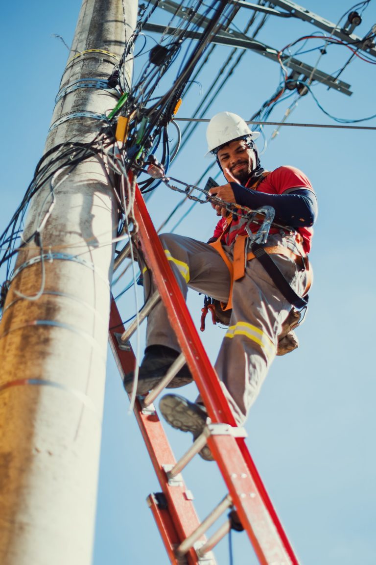 Electrician safely repairing electrical lines on a ladder under clear blue sky.