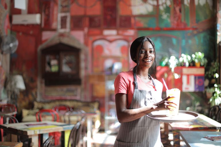 Happy waitress in apron serving a drink inside a vibrant, artistic cafe setting.