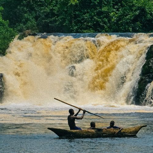 Three people canoeing near a powerful waterfall amidst lush greenery.