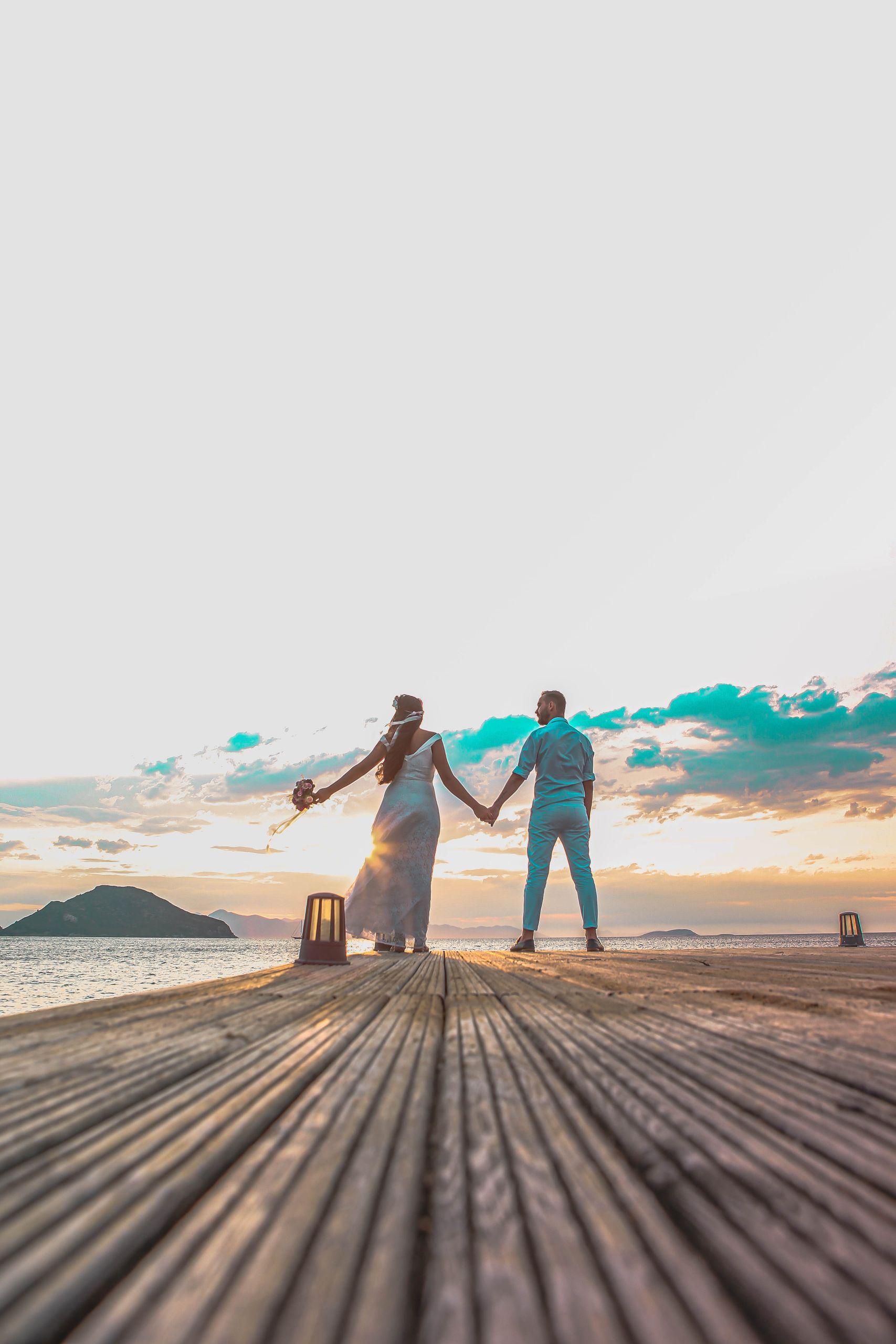 A romantic wedding couple holding hands on a wooden pier at sunset, capturing love and intimacy.
